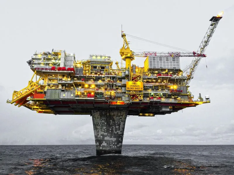 A large offshore oil platform stands isolated in the sea, supported by a tall concrete column. The platform is filled with intricate structures, pipes, cranes, and machinery painted in shades of yellow, red, and gray, contrasting against the cloudy sky and dark ocean waves. Lights illuminate the platform, highlighting its industrial complexity.